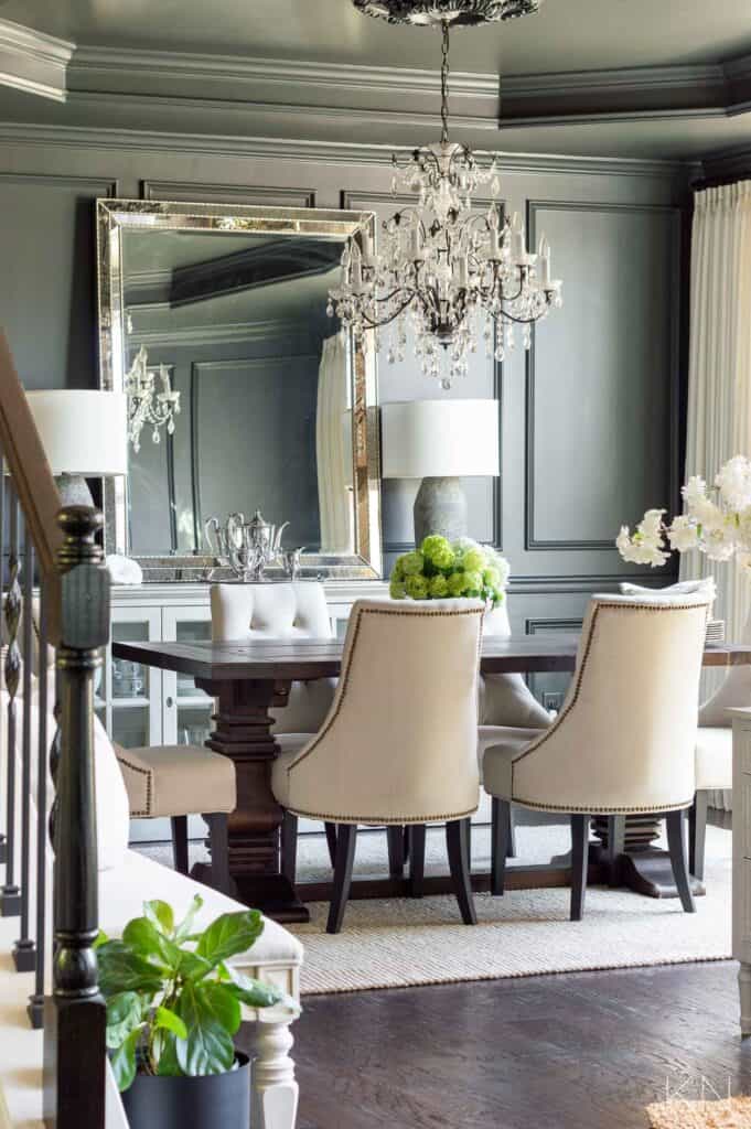 A dining room with dark walls and ceiling, a dark wood table and cream colored upholstered chairs with a fancy chandelier and a large mirror on a console table.