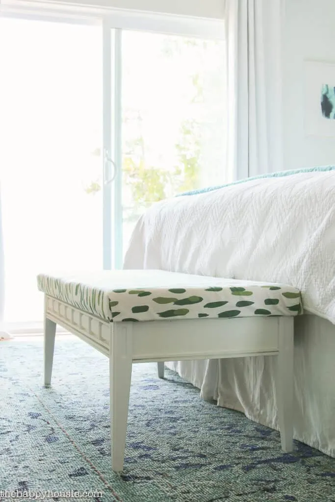 A white bench with white and green upholstered top at the foot of the bed with white bedding and a sliding glass door in the background.