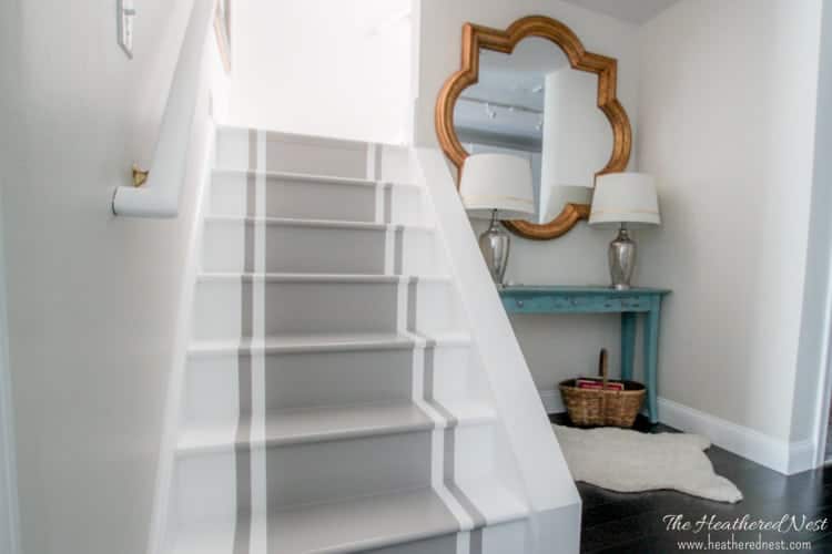 Light gray walls, white painted stairs and a gray painted runner down the center of the stairs with a light blue console table next to it with two silver lamps.