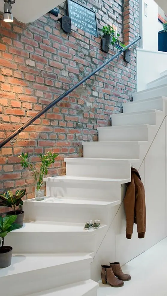 A white staircase with a brick wall, a black metal handrain, and a couple plants on the bottom few stairs.