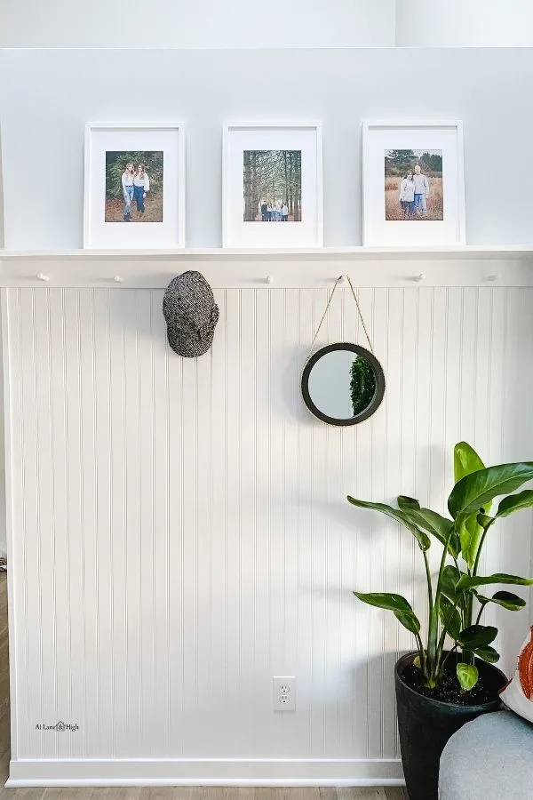 A white beadboard wall with a shelf holding family photos and pegs holding a hat and a round mirror.