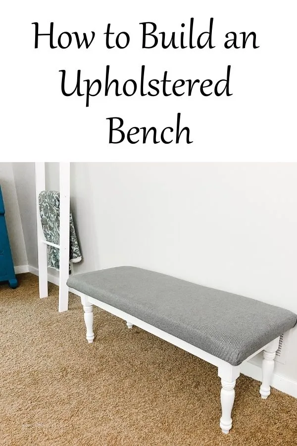 an upholstered bench with a white base and gray fabric on brown carpet next to a gray wall.