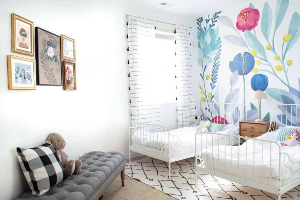 A girls bedroom with two iron beds, a floral mural on the headboard wall and striped curtains with tassels.