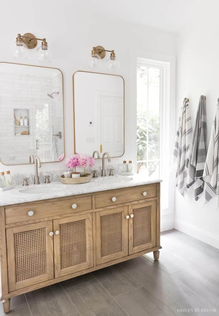 A bathroom with wood cabinetry, marble counters, gold hardware and two gold rounded edged rectangular mirrors.