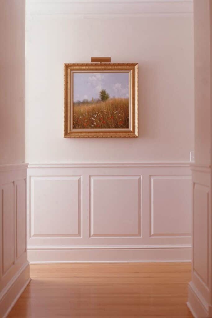 Raised panel wainscoting painted white with a creamy wall above and a landscape print with gold frame hanging on the wall.