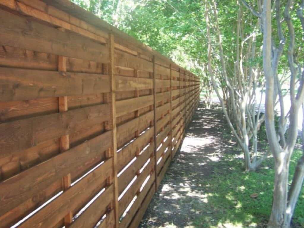 A horizontal fence with alternating slats and tall ornamental trees.