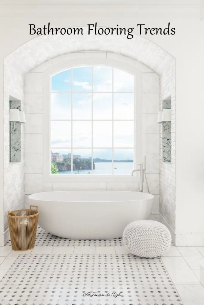 This bathroom has gorgeous white marble floors, a white free standing tub under a window overlooking the ocean, a basket and a pouf sitting next to the tub.