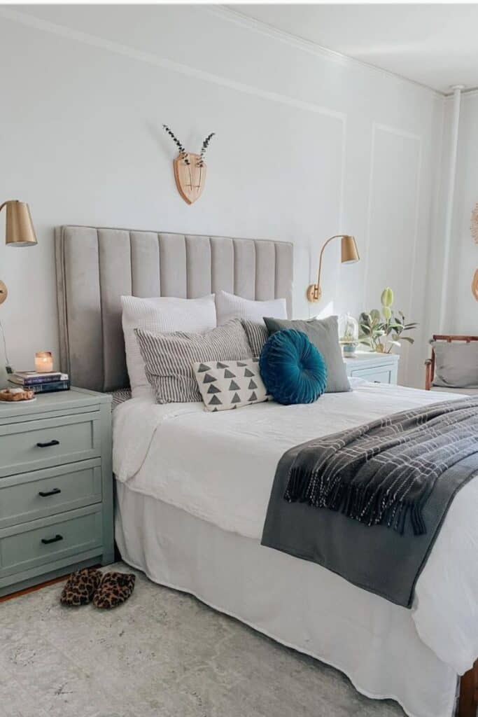 A bedroom with Nebulous White on the walls, white bedding and a blue green nightstand with gold lights attached to the wall above the nightstands.