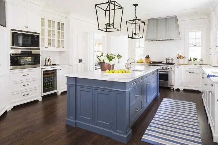 A mid toned blue island in a white kitchen, marble-looking counters and dark hardwood floors with a striped rug that matches the blue island.