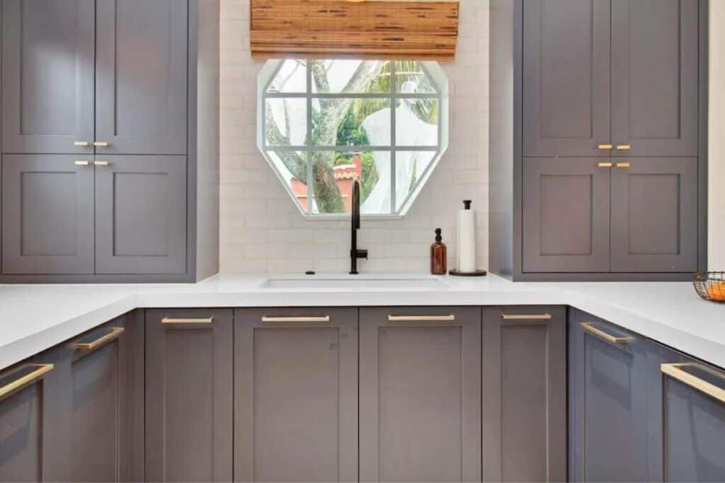 Dark Gray cabinets with gold hardware, white counters, and an octagon window over the sink.