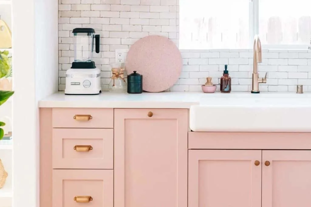 blush pink cabinets with a white counter and white farmhouse sink with gold hardware.