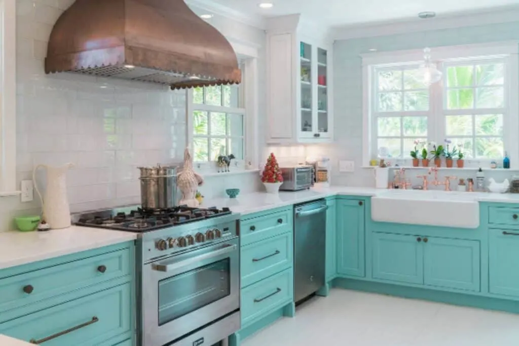 Turquoise cabinets with black hardware, white counters and backsplash with a copper oven hood vent.