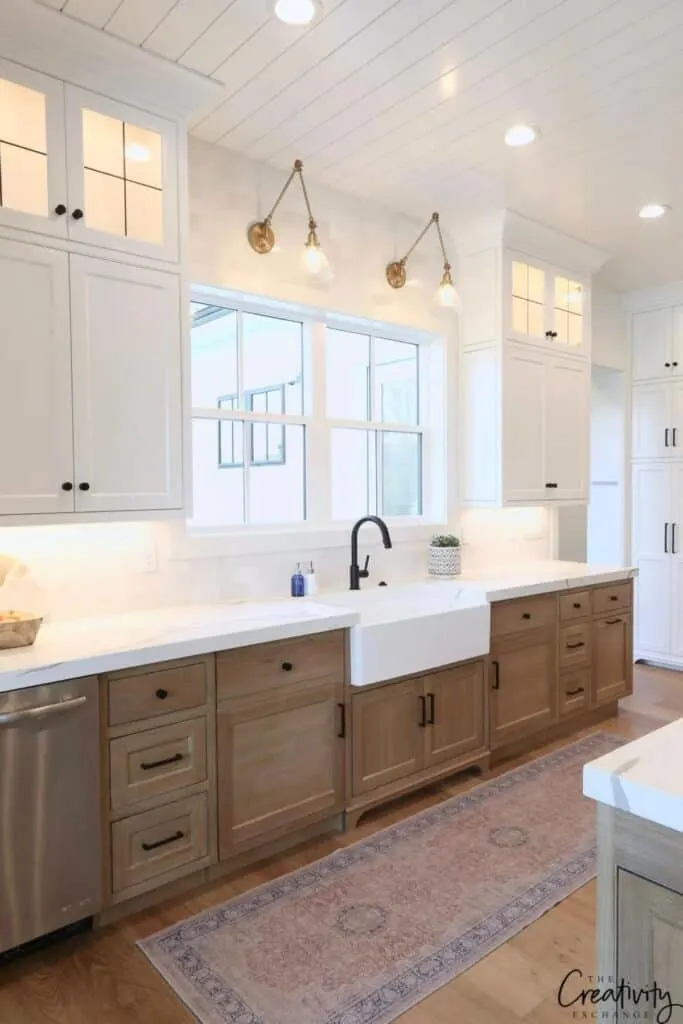Natural wood base cabinets with white uppers, large windows over the sink and a tongue and groove white ceiling.
