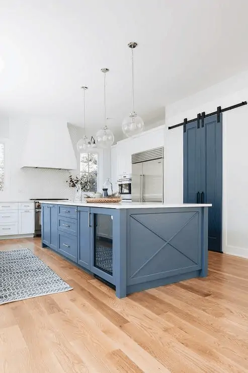 A white kitchen with light wood floors and a medium toned blue island with the same color in barn doors.