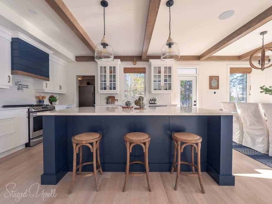A dark blue island in an all white kitchen with a dark blue stove hood and wood beams on the ceiling.