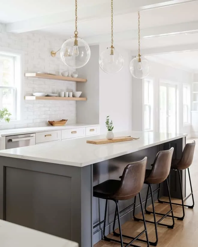 An all white kitchen with a very dark gray island and leather bar stools with light hardwood floors and floating shelves.