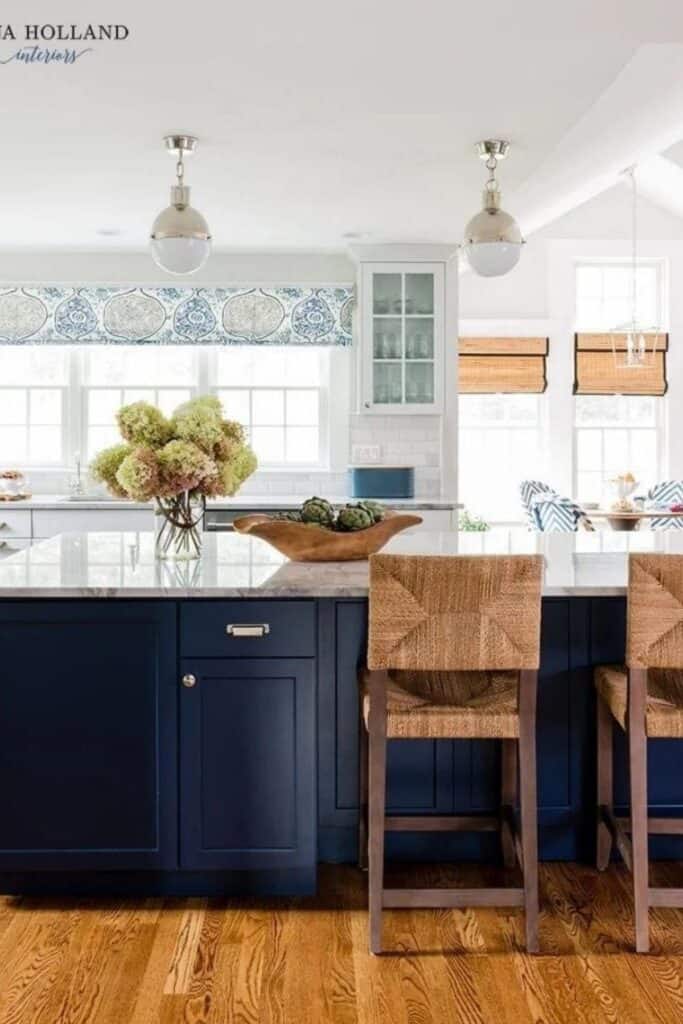 A dark blue island in an all white kitchen with wood bar stools and a vase of hydrangeas on the counter.