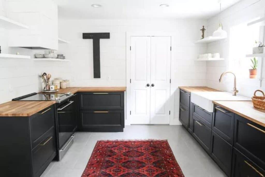 Black lower cabinets with butcher block counters, white shiplap walls and a dark red geometric rug in the center of the floor.