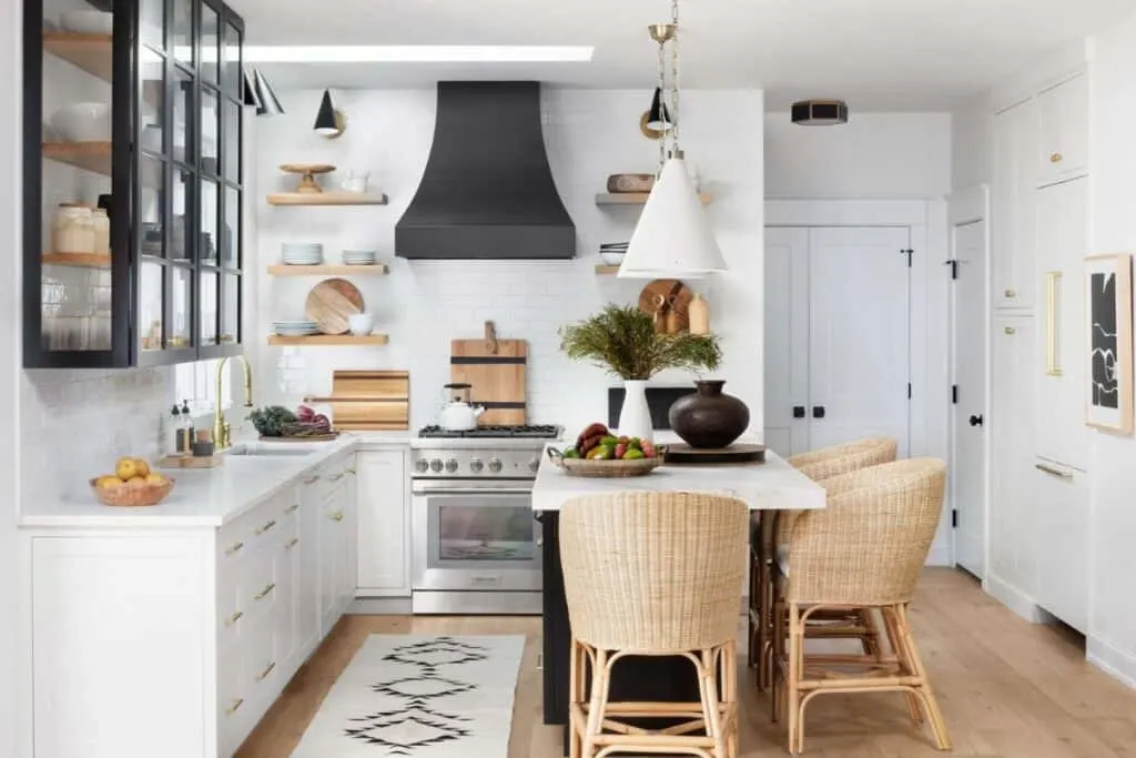 A white kitchen with a black oven hood and island, light rattan bar stools and wood accents in the accessories.
