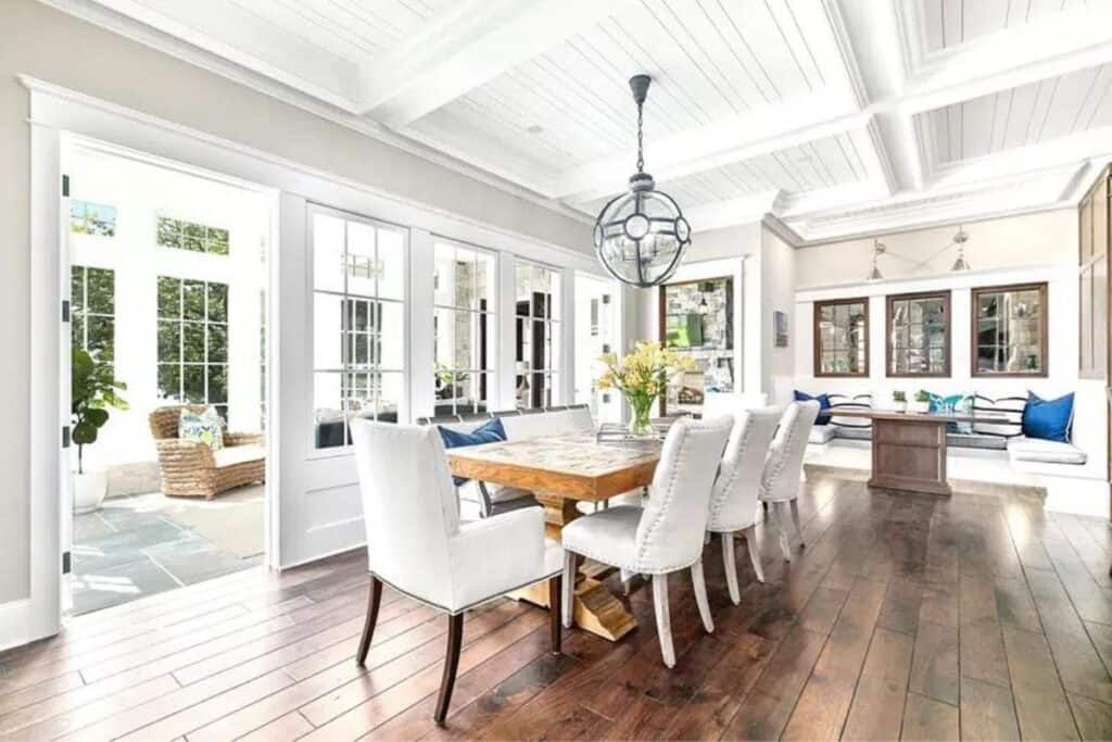 A dining room with rustic wood floors and table, white upholstered chairs and a coffered ceiling with beadboard on the ceiling panels.