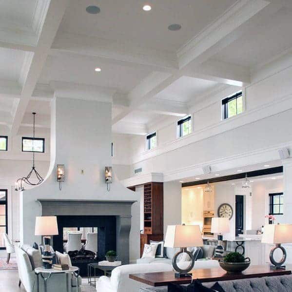 An all white family room with a huge fireplace between the family room and dining room and tall ceilings that are coffered.