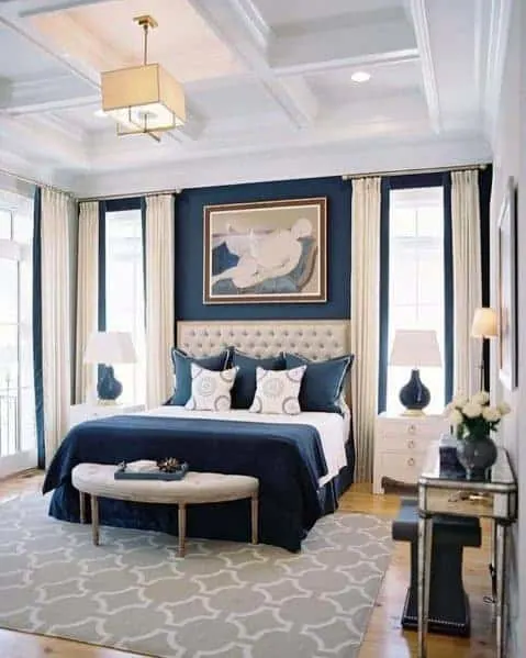 A bedroom with navy walls, cream headboard and curtains with a white coffered ceiling.