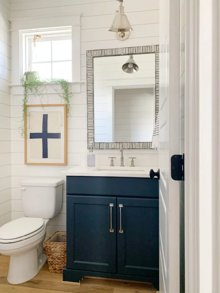 A powder room with Hale Navy on the vanity cabinet, shiplap on the walls painted white, a maritime cross flag framed on the wall and a plant hanging above.