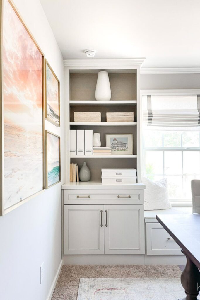 An office with On The Rocks on The walls, blue and coral wall art, shelves with white and beige accessories and a dark stained table for a desk.