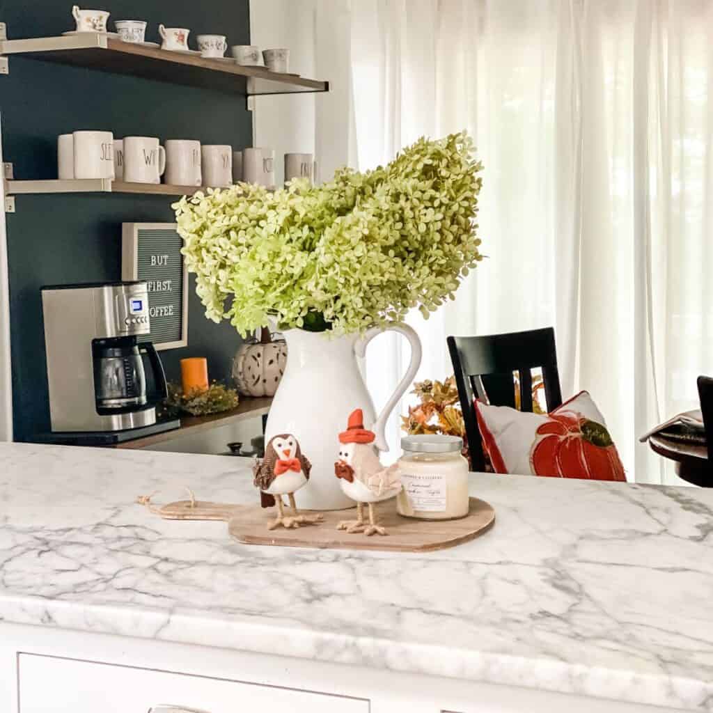 A vignette with a pitcher of hydrangea flowers, two stuffed birds and a candle on a cutting board on my kitchen counter with the breakfast nook in the background.