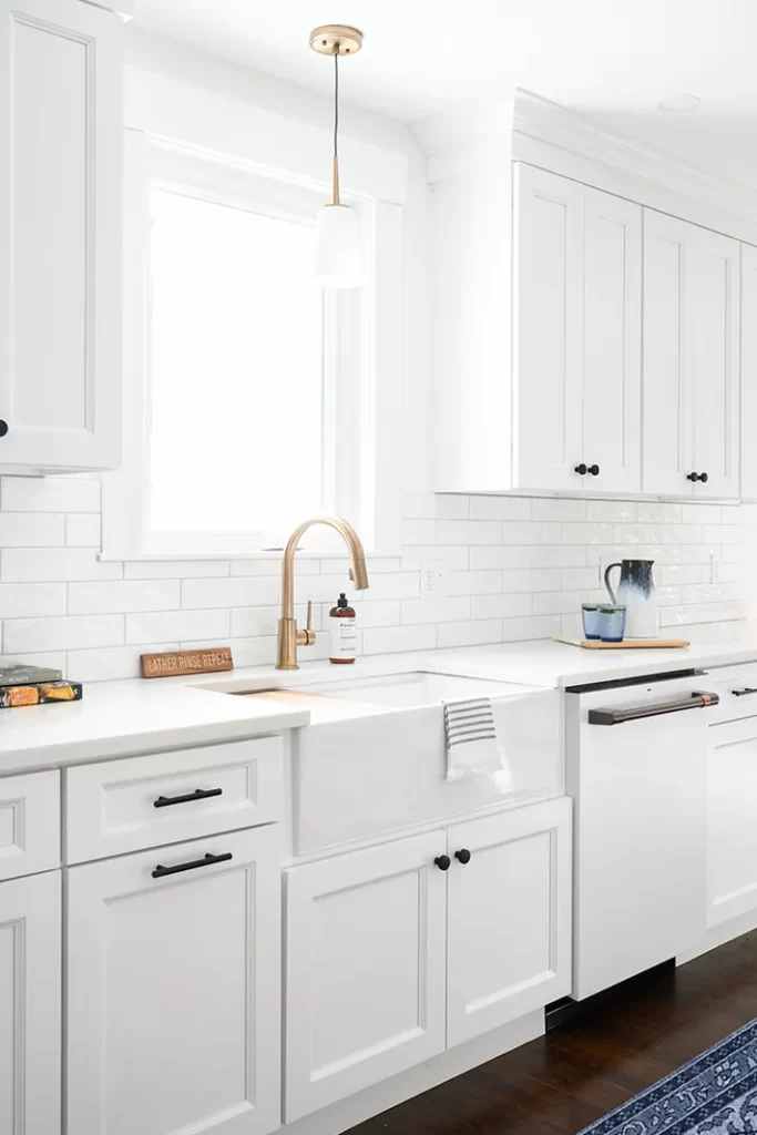 White kitchen with white subway tile backsplash, white counters, white farmhouse sink and gold faucet.