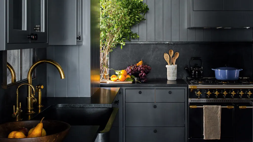 An all black kitchen with gold hardware, tall green stems in a vase and lots of fruit on the counter.