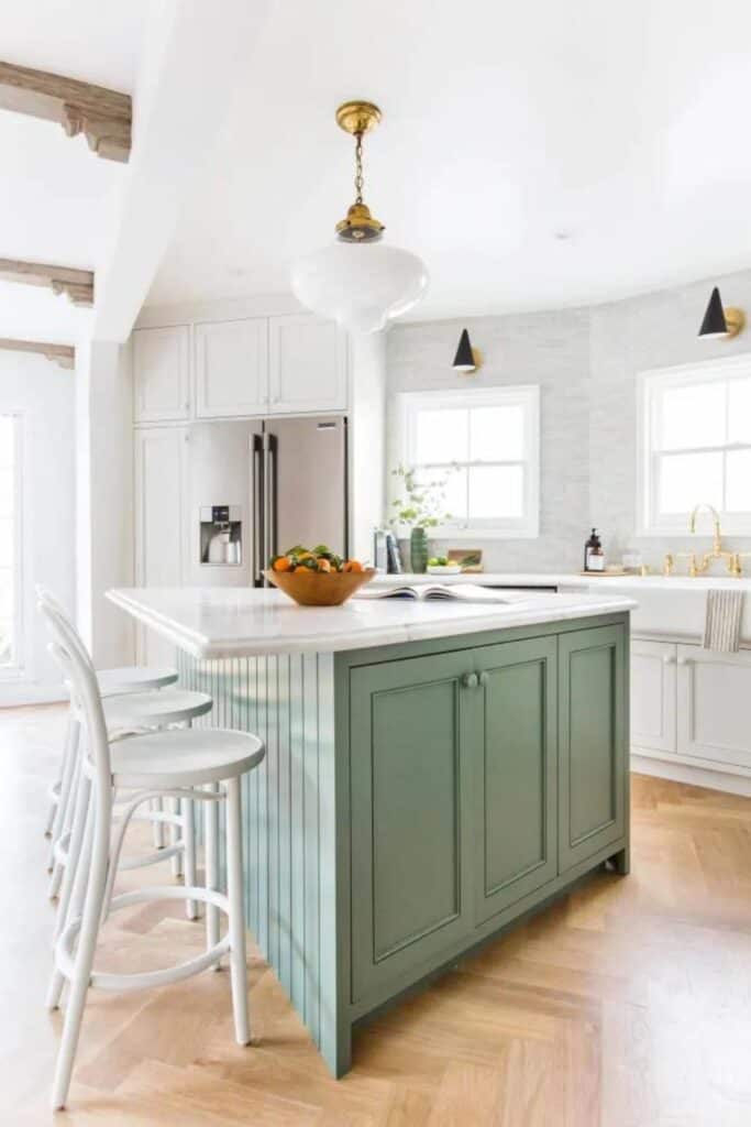 A kitchen with white outer cabinets and a sage green island with white countertops and herringbone floors.