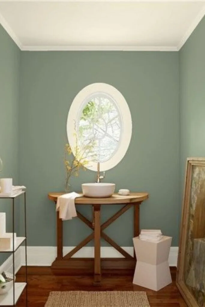 A bathroom with Sage Wisdom painted on the walls, a wood vanity and vessel sink with an oval window above.