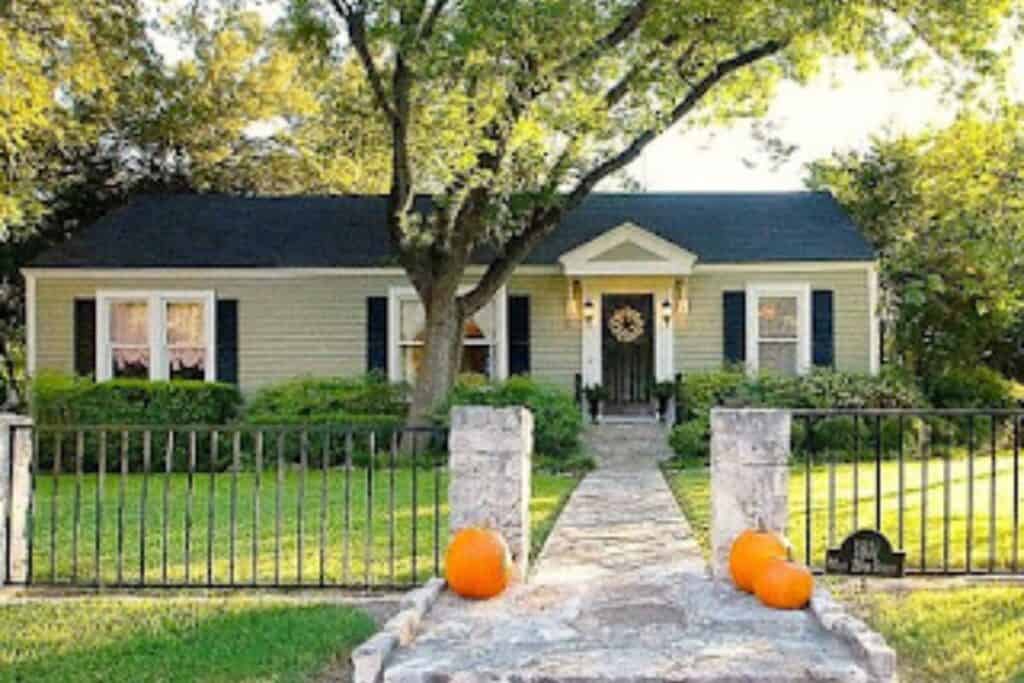 The exterior of a ranch home with Texas Sage, black door and shutters with a sidewalk flanked by pupkins.
