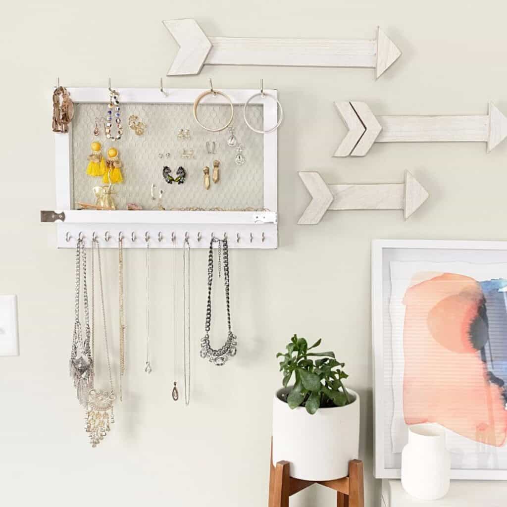 A wood wall mounted jewelry holder painted white with hooks at the top and bottom and chicken wire in the middle to hang earrings.