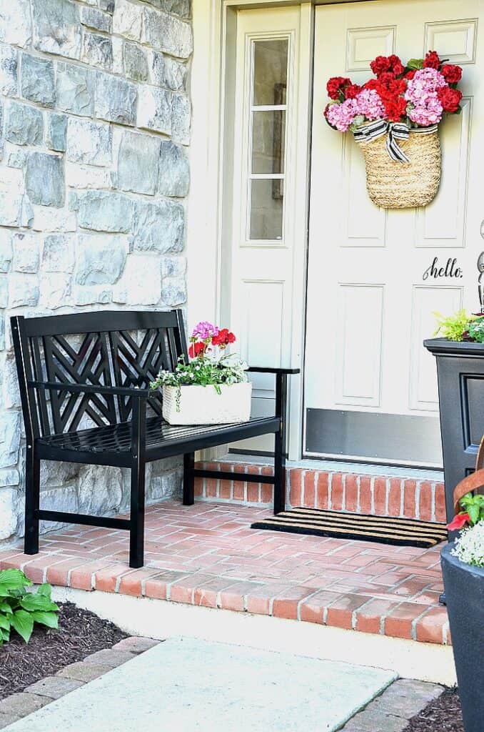 A small front porch iwth a black iron bench, a small planter on the bench and a basket of flowers hanging on the door.