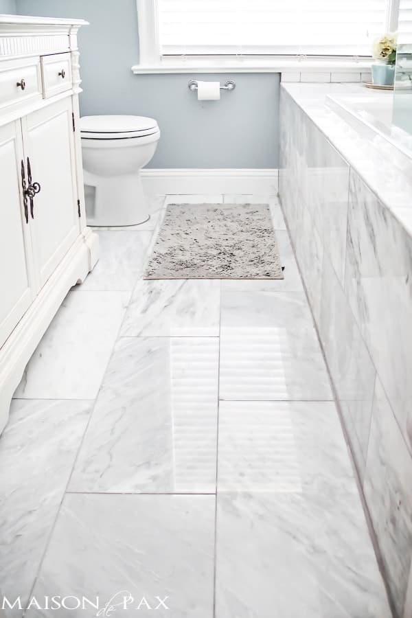 A beautiful bathroom with blue gray paint color on the walls, marble floors, white cabinets and white subway tile on the tub.