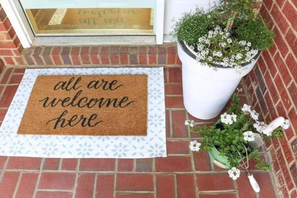 A small front porch of a red brick home with layered mats that says all are welcome here and two plants that have white flowers in them.
