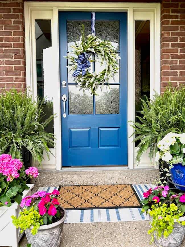A small front porch with a bright blue door, layered door mats and lots of brightly colored plants.