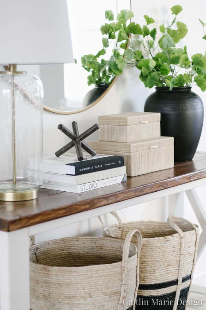 A console table with a round mirror above, black vase with greenery, clear lamp and books with a large black jack.