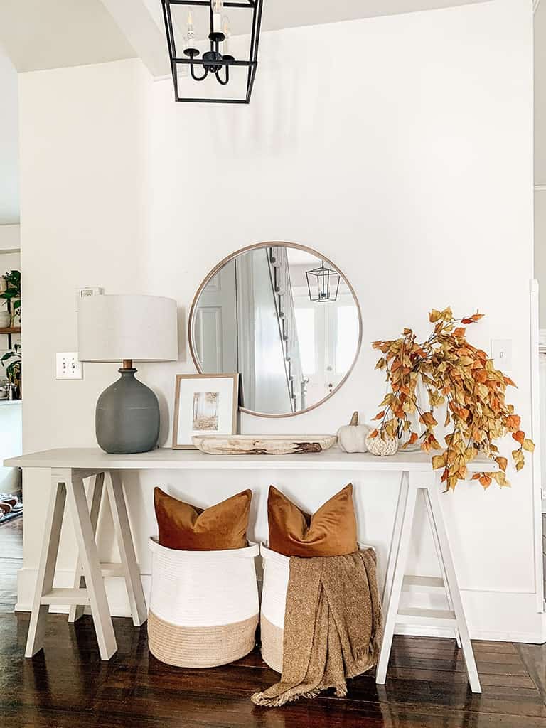 A long console table with a gray lamp, round mirror, and baskets below holding pillows and blankets.