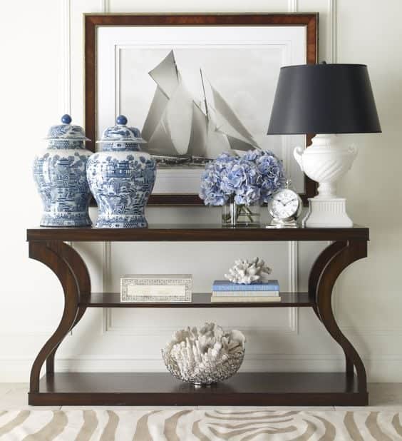 A dark wood console table with curved ends, a print with a sailboat on it, a white lamp with a black shade and two blue and white ginger jars.