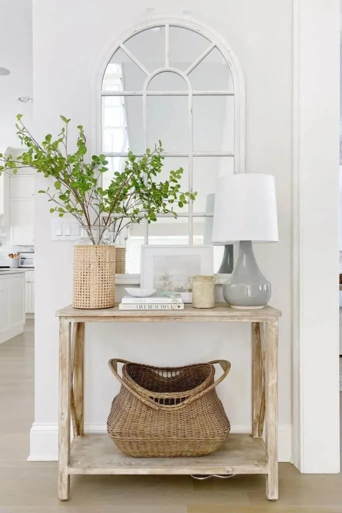 A light wood console table with a large arched mirror above, a gray lamp with a white shade, and a vase with leafy branches.