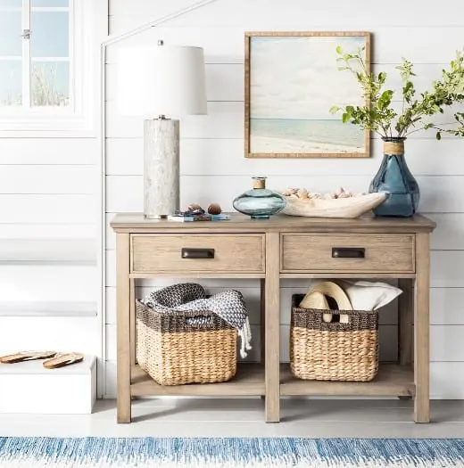 A light wood console table with a coastal print above, white tall lamp and driftwood bowl.