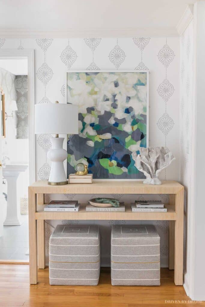 A light wood console table with two gray pinstripe ottomans below, a blue, gray, and black art print above, a white lamp and books on top.