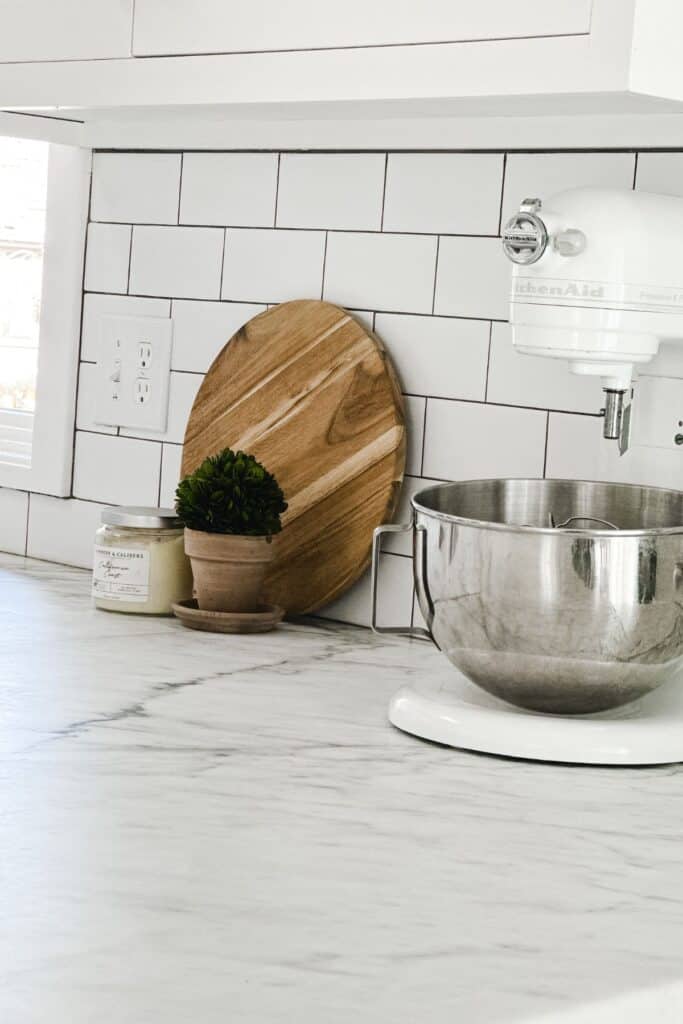 White marble counters with a KitchenAid mixer, white subway tile backsplash and a cutting board, candle and plant in front of it.