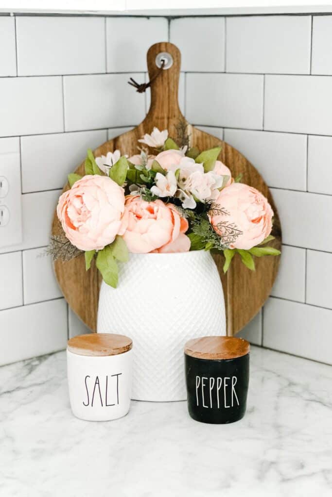 A vase with pink peonies, a salt and pepper shaker with a round cuttingboard behind them against a white subway tile backsplash.