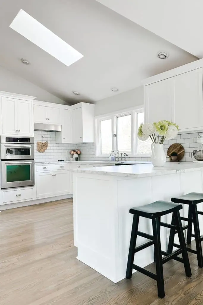A wide view of a kiitchen with white cabinets, stainless appliances, black barstools and light hardwood floors.