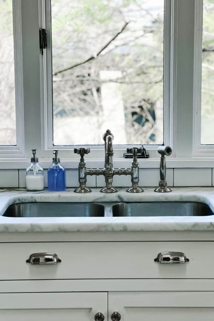 A view of a sink with whtie cabinets, marble counters, stainless faucet and large windows above.