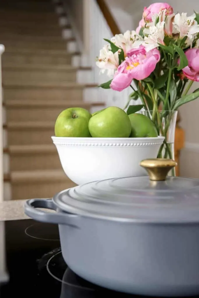 A stovetop with a gray dutch oven, a white pedestal bowl with green apples and a vase with pink flowers.